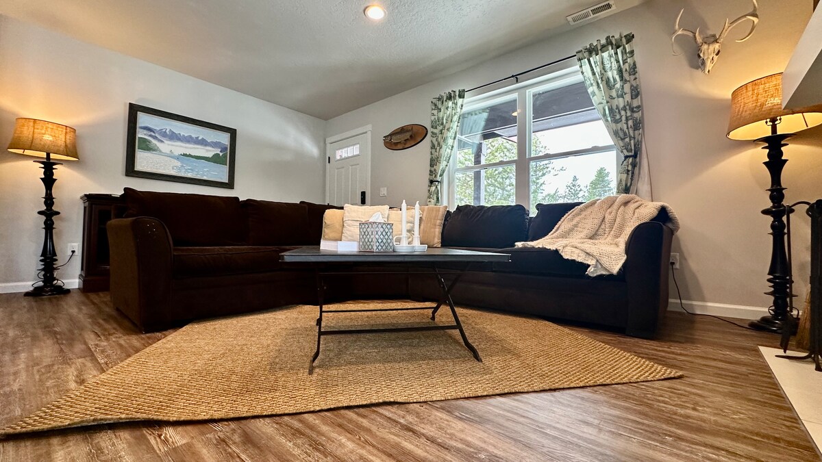 A spacious living area features a dark brown sectional sofa, complemented by decorative pillows and a soft throw blanket. A coffee table sits centrally on a woven rug. Two standing lamps provide soft lighting beside the windows, which are dressed with patterned curtains.