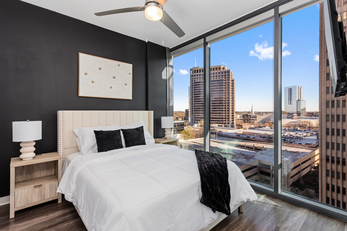 A cozy bedroom features a spacious bed with white bedding, flanked by two table lamps on nightstands. A large window offers a city view, allowing natural light to fill the room. A ceiling fan is visible, enhancing the modern look of the space.