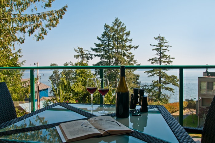 A glass balcony table is set with two wine glasses, a bottle of wine, and a book, overlooking a serene waterfront view. Lush green trees can be seen in the foreground with the ocean in the background, creating a calming atmosphere.