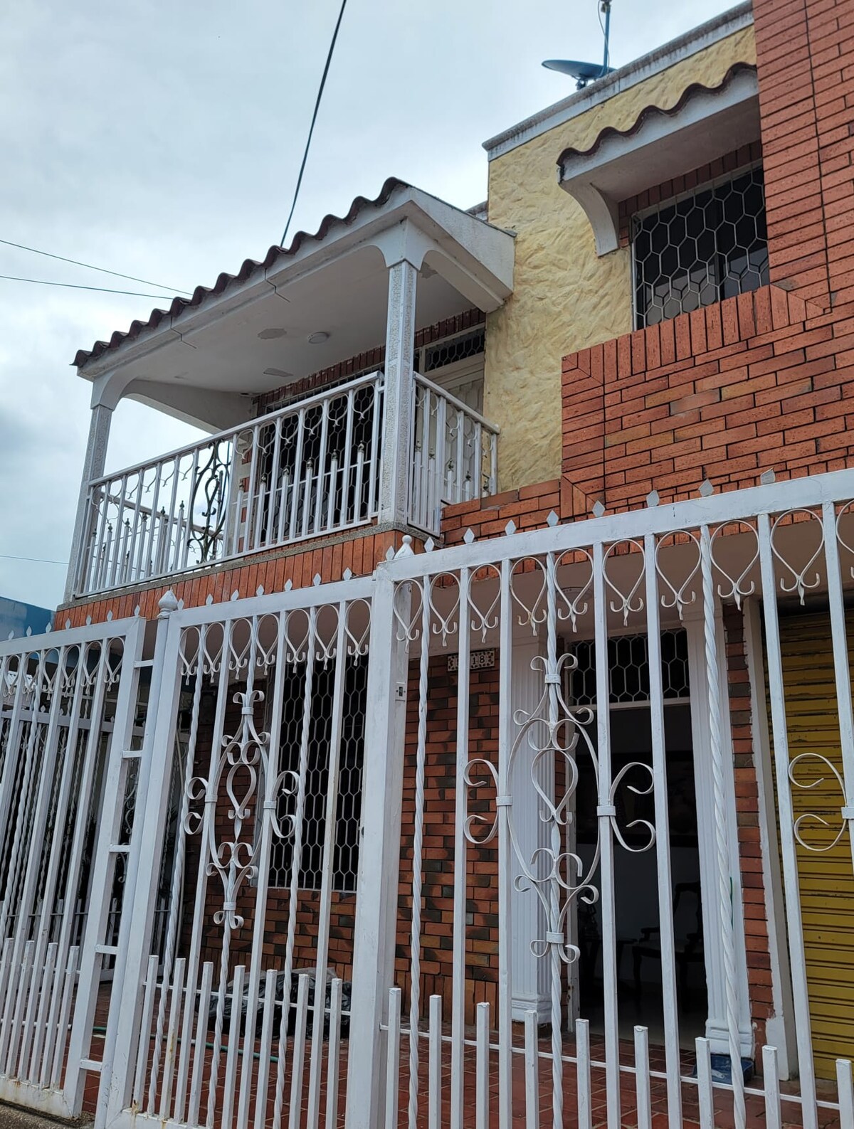 The exterior of the house features a combination of yellow and red brick, framed by a white wrought iron fence. A balcony with decorative railings is visible above the entrance, contributing to the architectural charm of the property.