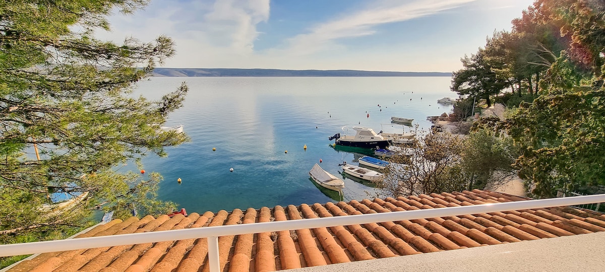 A serene view showcases calm waters reflecting the sky, with several boats anchored nearby. Pine trees frame the image, adding natural elements to the scene. A terracotta-tiled roof leads the eye towards the tranquil waterfront.