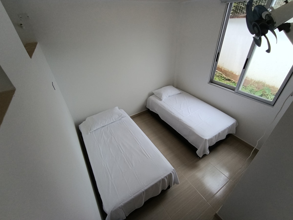 A bright room features two twin beds neatly made with white linens. Natural light enters through a large window, illuminating the simple decor. The floor is tiled, and minimal furnishings enhance the spacious feel of the area.