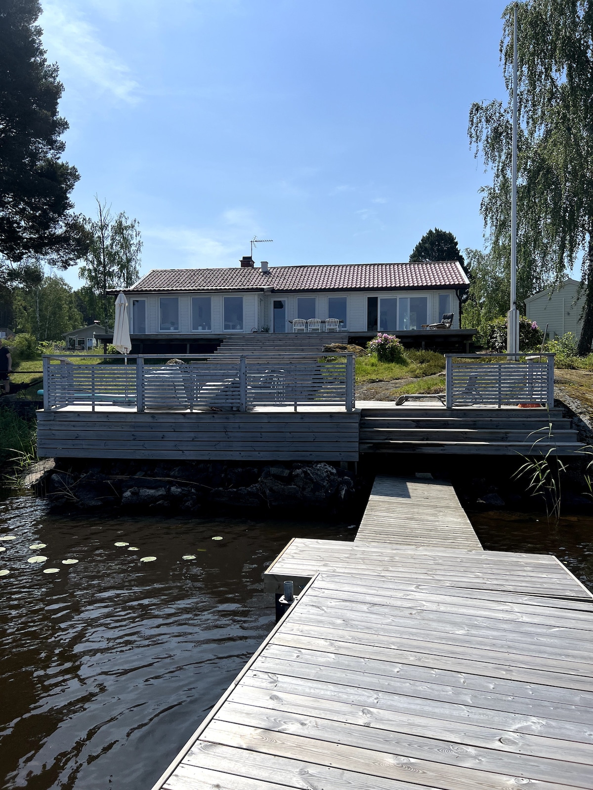 The exterior of the property is visible, featuring a wooden deck that leads to a private dock. The main building has large windows, allowing for ample natural light. Greenery surrounds the area, with a clear blue sky overhead, creating a serene environment.