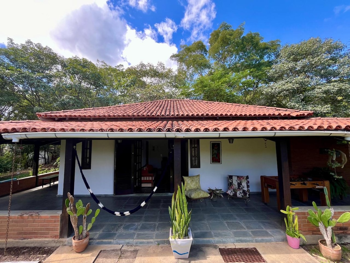 A charming one-story structure features a traditional red-tiled roof and a stone pathway leading to the entrance. Potted plants are positioned in front, while the surrounding greenery creates a serene backdrop. Large windows allow ample natural light into the interior.
