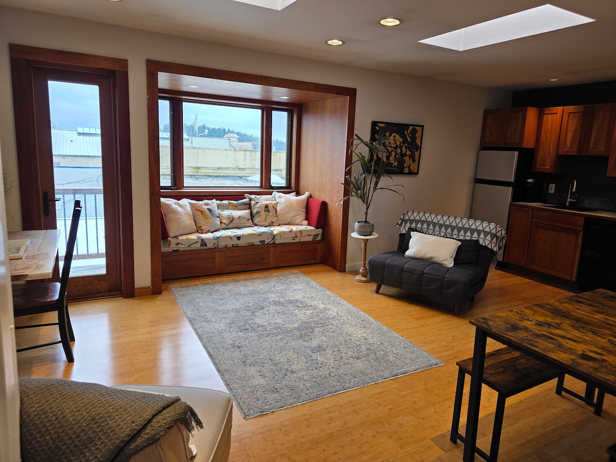 A cozy corner nook is highlighted by a built-in bay window seat adorned with colorful cushions, offering views of the outdoors. A dining area is visible with a wooden table and chairs, complemented by bamboo flooring, while a well-equipped kitchen is seen in the background.