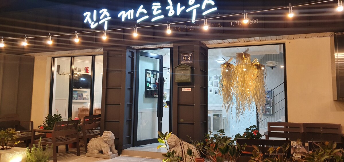 The entrance of the guesthouse is framed by strings of lights, creating a welcoming ambiance. Two decorative stone figures sit on either side of the entrance, complemented by planters filled with greenery. Large glass doors provide a view into the lobby area.