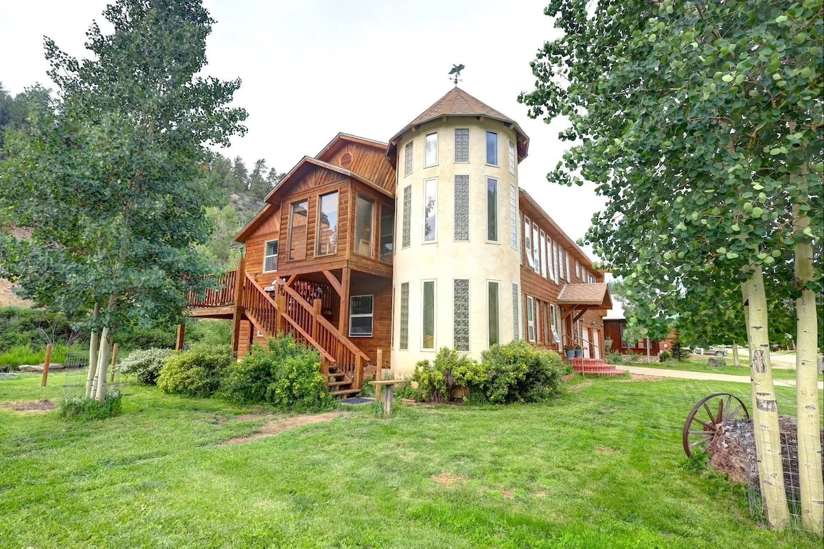 A unique two-story structure features a circular tower and a large rectangular section, both adorned with multiple windows. Surrounded by a lush green lawn, the building is set against a backdrop of trees and gentle hills.