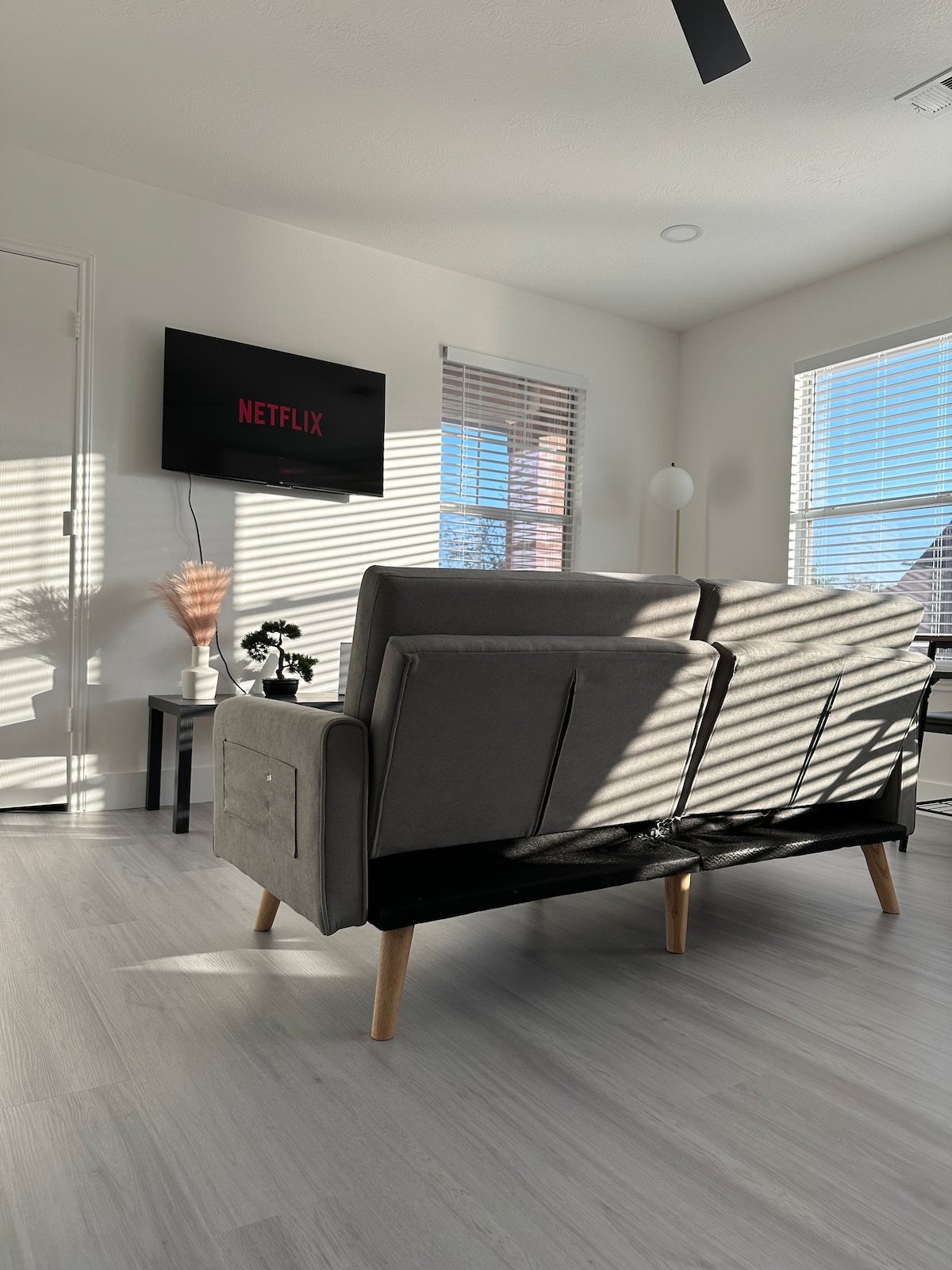 The living area features a modern sofa, oriented towards a wall-mounted smart TV displaying the Netflix logo. Natural light filters through window blinds, creating striped patterns on the light-colored flooring, complemented by a multi-functional side table adorned with a small plant and decorative elements.