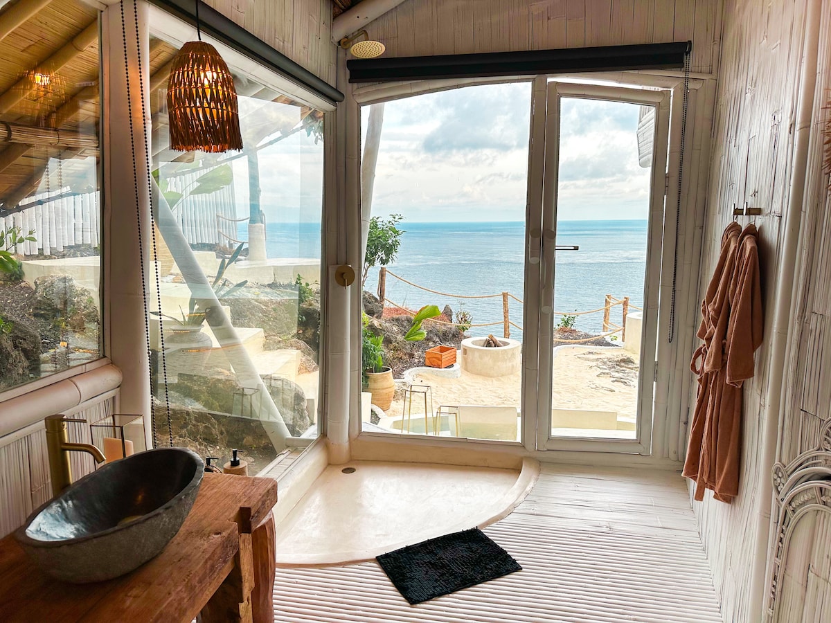 A bathroom is situated near large glass doors that offer views of the ocean. A wooden table, featuring a round sink, provides a natural touch. The space is complemented by a soft rug, and plush bathrobes hang invitingly nearby.