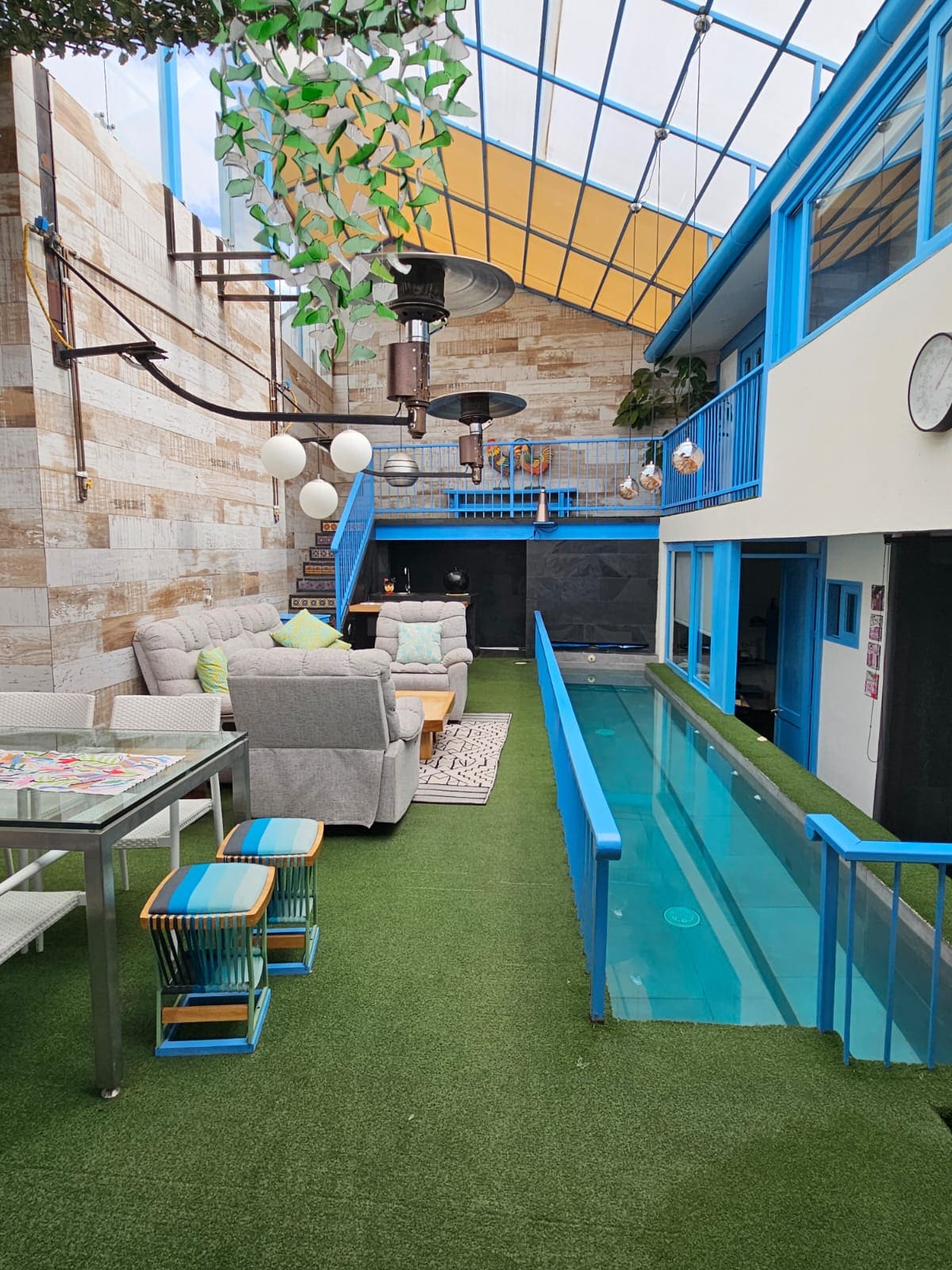 A modern living area features a sleek design with a clear wall pool running alongside. Comfortable seating is arranged around a glass table, with greenery adorning the space. Large windows allow natural light to fill the room, enhancing the lively atmosphere.