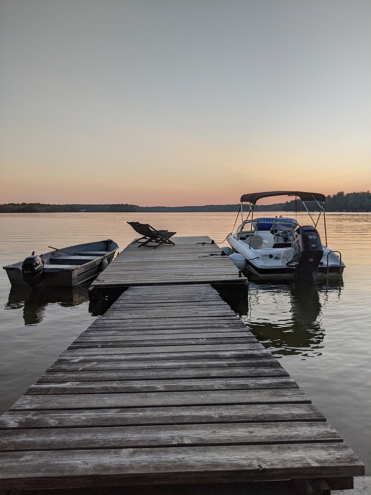 A wooden dock extends over the calm water, featuring two boats moored alongside. A single lounge chair is positioned on the dock, facing towards the peaceful water at sunset, creating a serene and inviting setting.