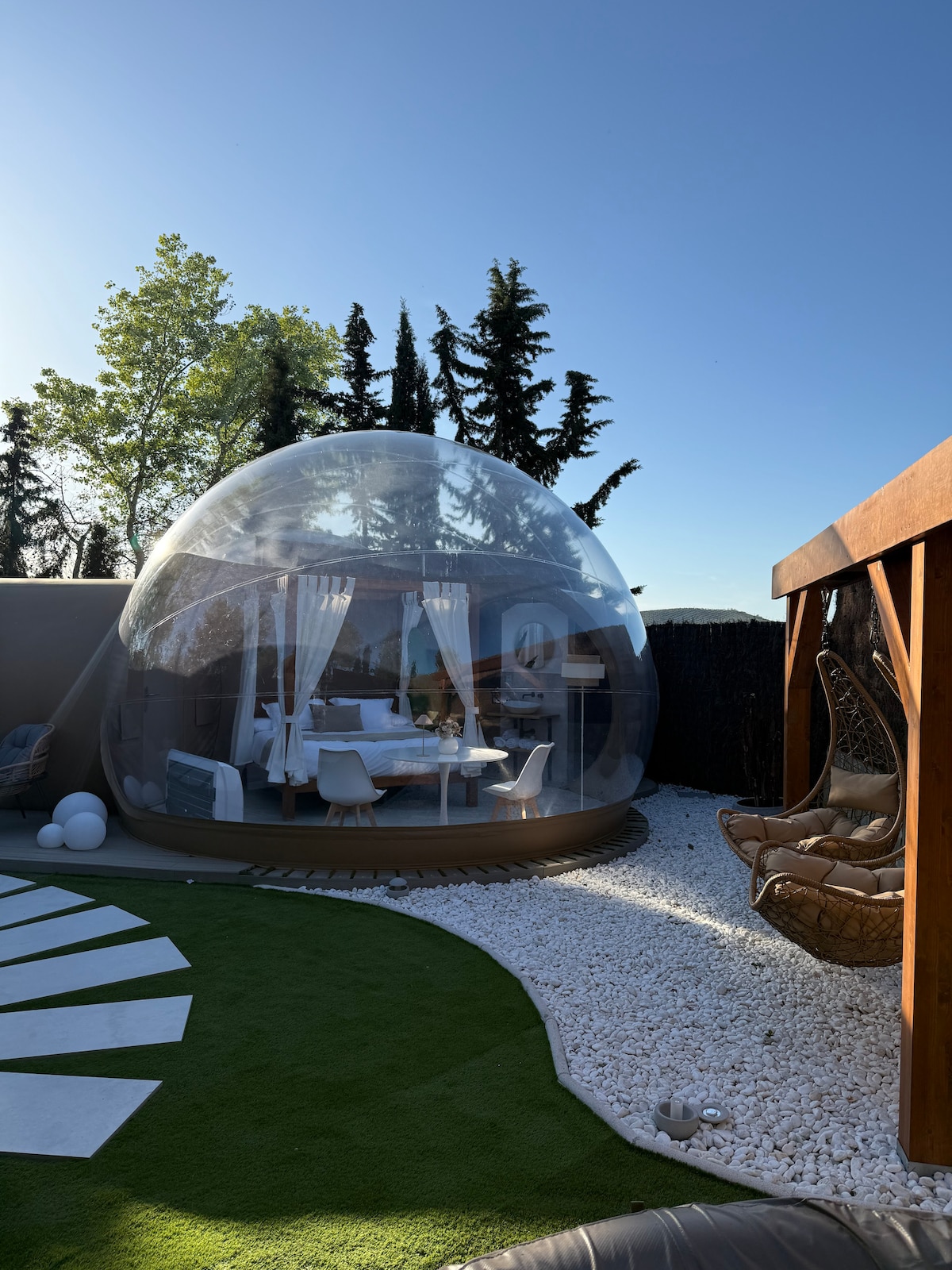 A transparent bubble structure is set within a landscaped area featuring green grass and white stones. Surrounding trees provide shade, while cozy seating is arranged under a wooden pergola. The interior of the bubble shows a bright and airy space with soft furnishings.