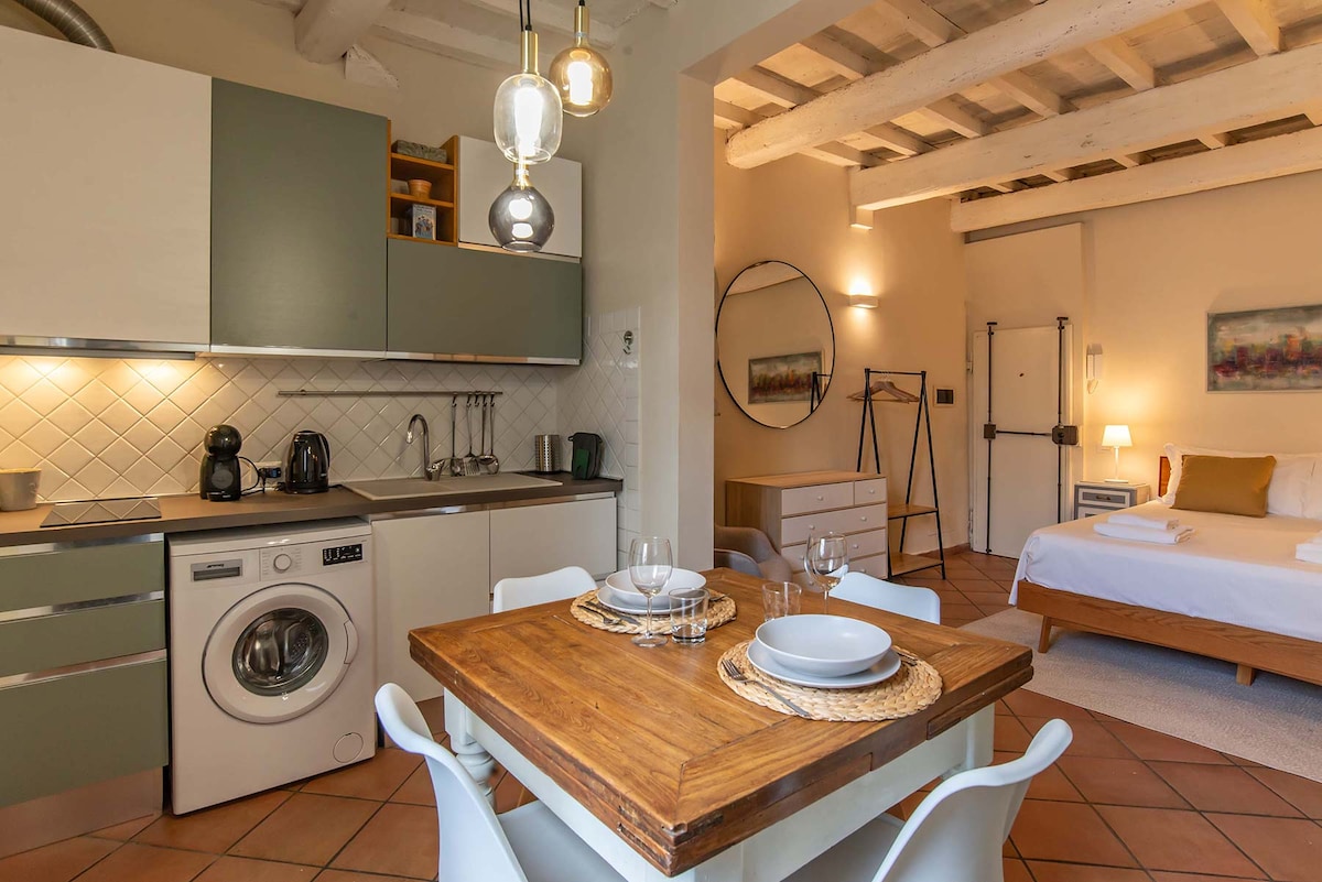 An open-concept living area features a wooden dining table set for four, complemented by modern white chairs. A well-equipped kitchenette is clearly visible, with appliances including a coffee maker and washing machine. Soft lighting highlights the natural textures of the room, and a spacious bed is positioned nearby.