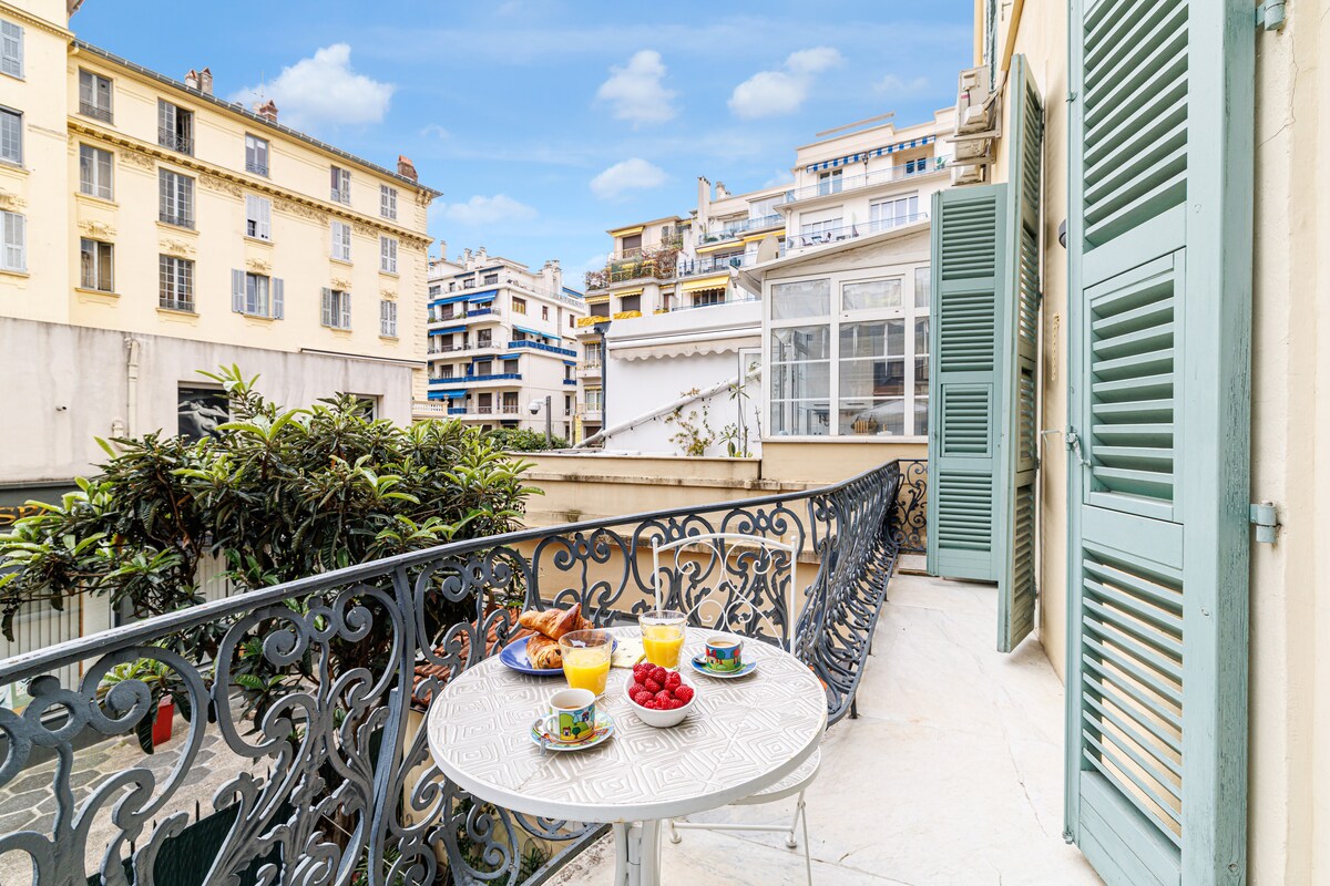 A charming balcony is presented, featuring a small round table set for breakfast with fresh pastries, fruits, and two glasses of juice. Elegant wrought iron railing frames the view, while large windows with shutters provide natural light and an inviting atmosphere.