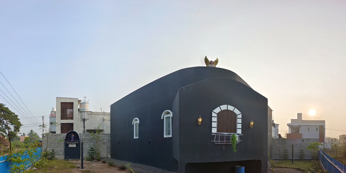 A unique dome-shaped structure stands prominently against a serene sunrise backdrop. The exterior features dark walls with contrasting white accents, including arched windows and a decorative entryway. Lush greenery surrounds the property, enhancing the tranquil atmosphere.