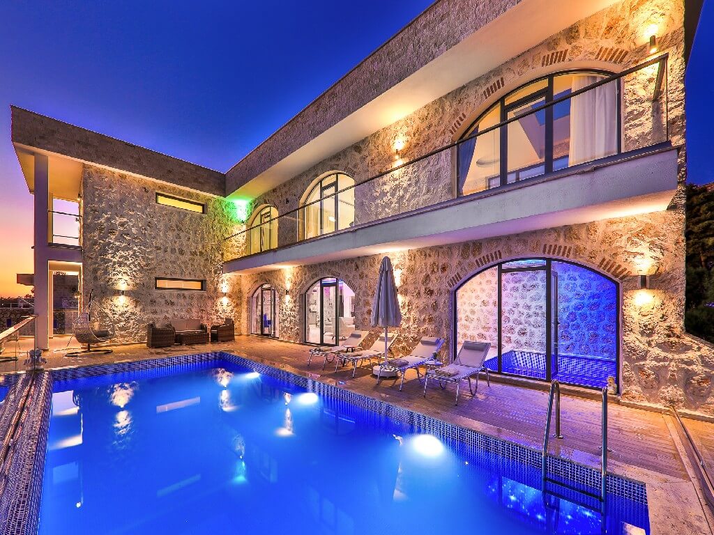 A stone villa is illuminated by soft lighting at dusk, showcasing a large outdoor pool. Sun loungers are arranged around the pool, and spacious glass doors provide access to the interior. The building features multiple balconies with elegant arches and large windows.