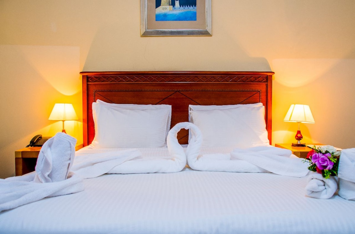 A cozy room featuring a spacious bed adorned with neatly arranged white linens. Two bedside lamps cast a warm glow alongside a floral arrangement. A decorative painting is displayed above the headboard, contributing to a serene atmosphere.