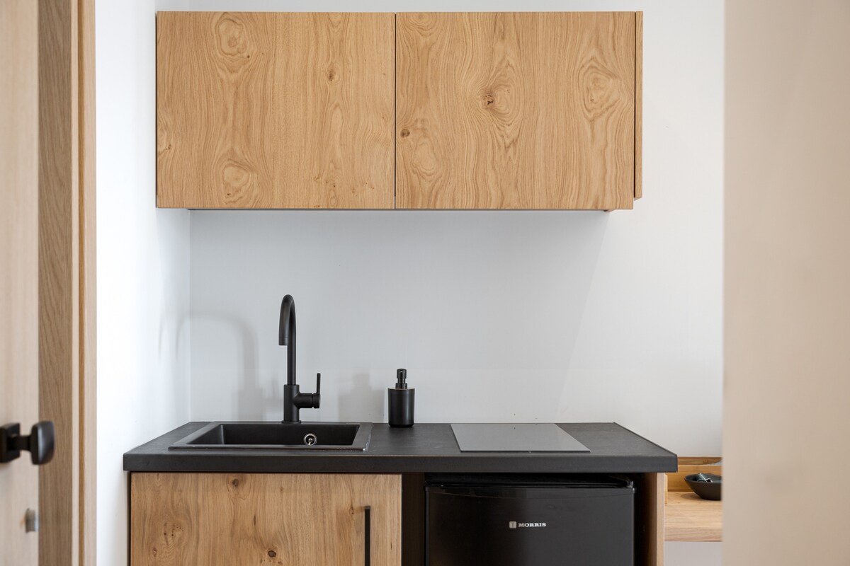 A compact kitchenette features modern wood cabinetry with a warm finish. A sleek black counter includes a sink and a single-handle faucet. An under-counter refrigerator is positioned underneath, and a small cooking surface is visible, designed for practical use in the space.