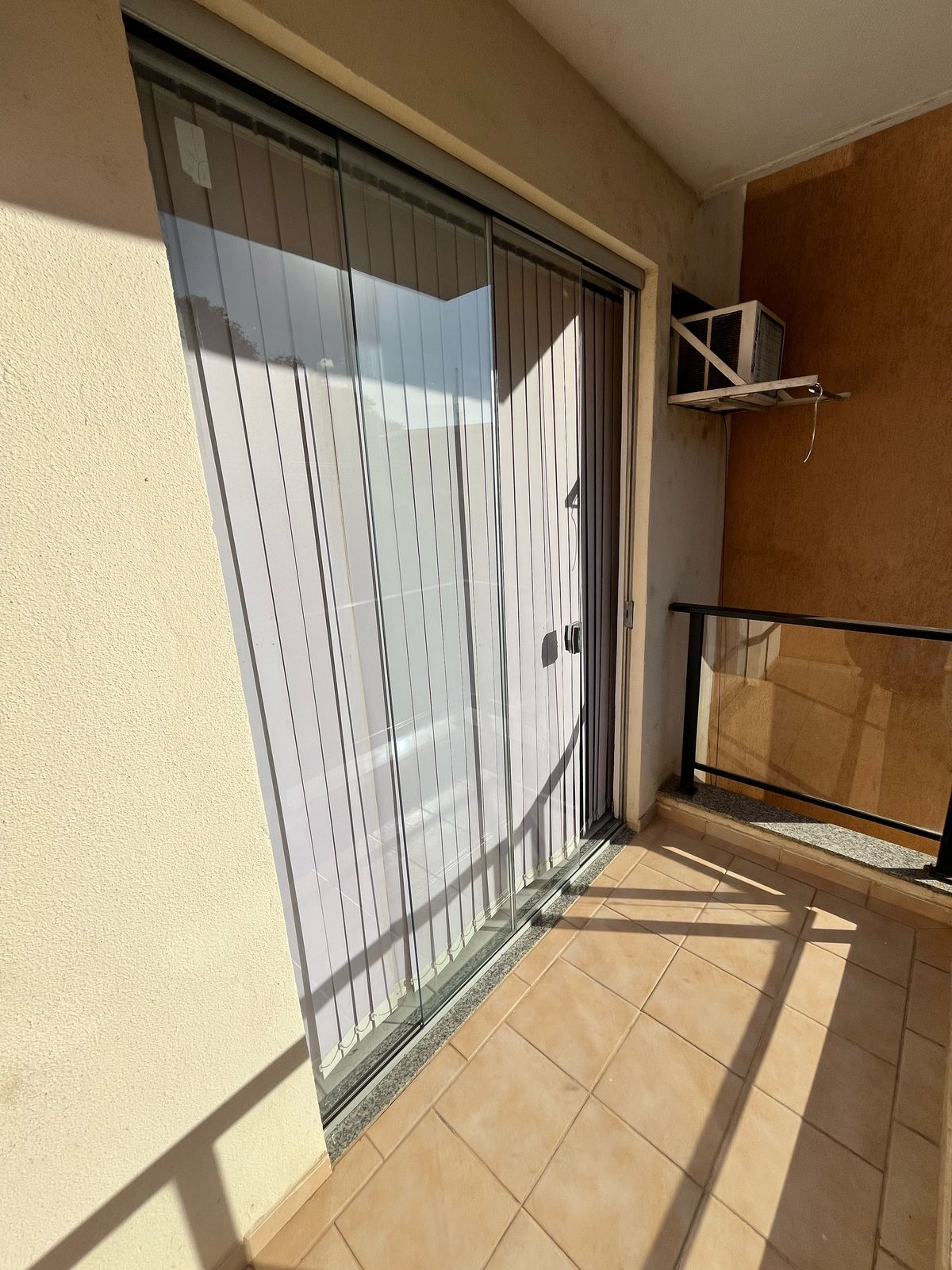 A balcony area is featured, showcasing large sliding glass doors with vertical blinds. The space is filled with natural light, highlighting tiled flooring. An air conditioning unit is mounted on the wall, and a railing surrounds the balcony for added safety.