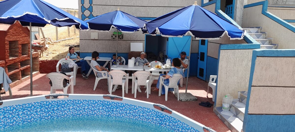 A spacious outdoor area is highlighted by a circular pool surrounded by blue patterned tiles. Several white chairs and a large table are placed under large blue umbrellas, providing shade. The building's exterior features blue accents and stairs leading to a welcoming entrance.