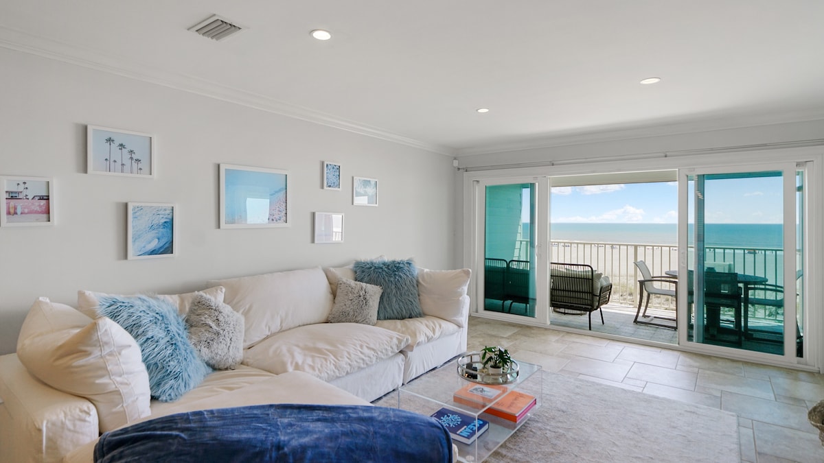 A bright living room features a spacious sectional couch adorned with soft blue and white pillows. Large glass doors provide access to the oceanfront balcony, with a panoramic view of the Atlantic. Artwork depicting coastal scenes adorns the light-colored walls.