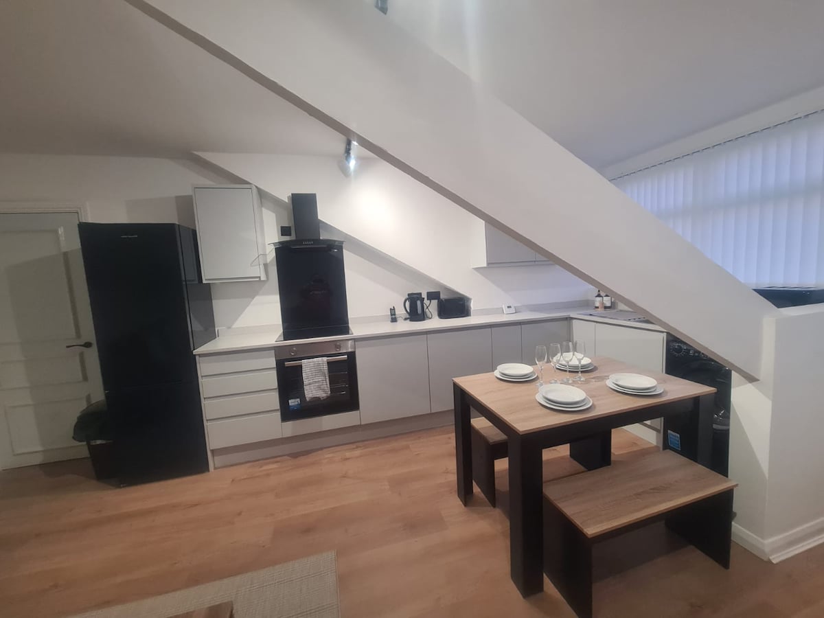 A modern kitchen is visible, featuring sleek cabinetry and a black refrigerator. A dining table set for four is positioned centrally, complemented by minimalist tableware. Natural light filters through the window, enhancing the warm tones of the wood flooring and creating an inviting atmosphere.