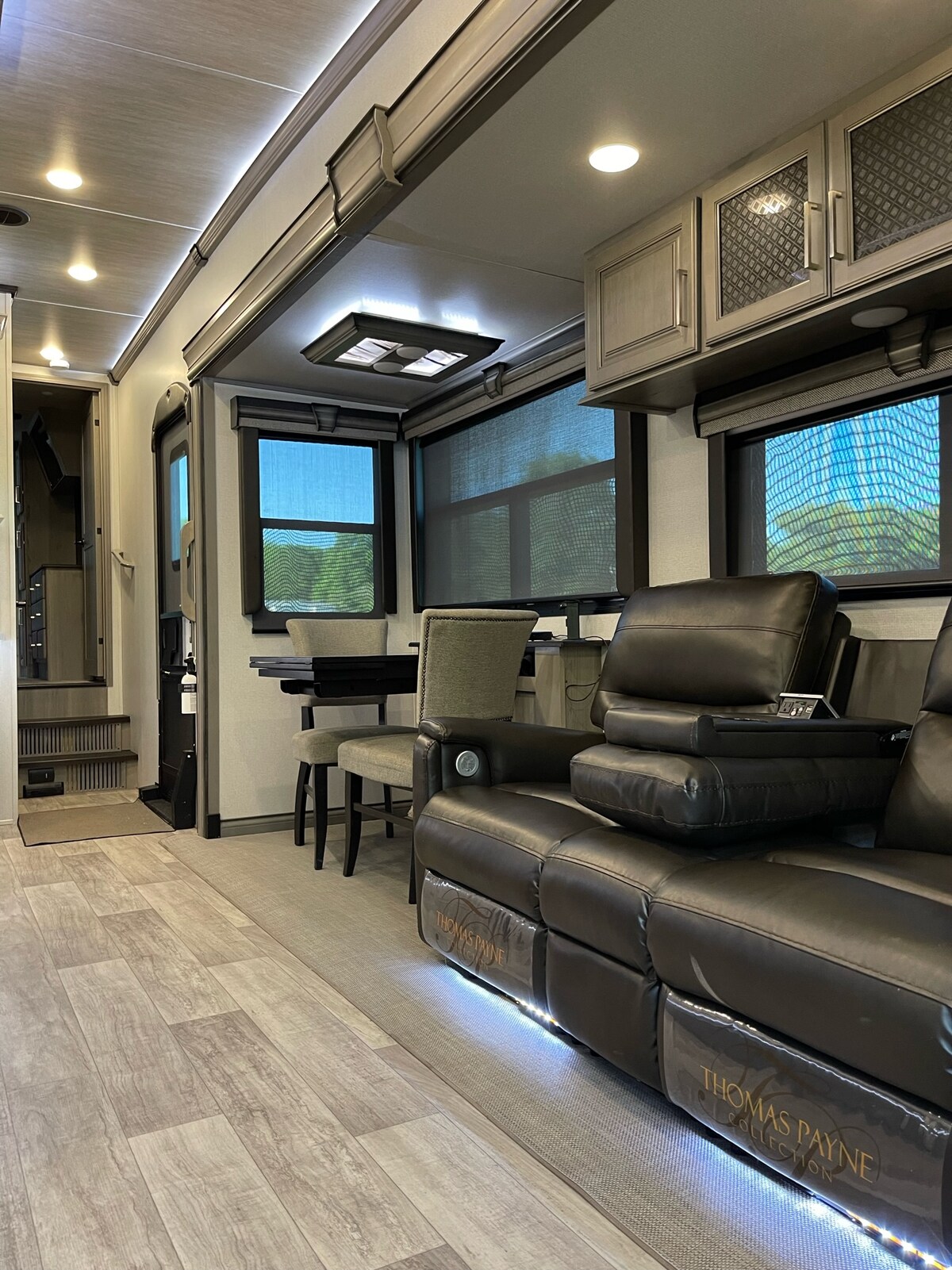A comfortable seating area features black reclining chairs with ambient lighting beneath. Large windows provide natural light, and a dining space with upholstered chairs is visible in the background. The modern design incorporates sleek cabinetry and a clean layout, enhancing the cozy ambiance.