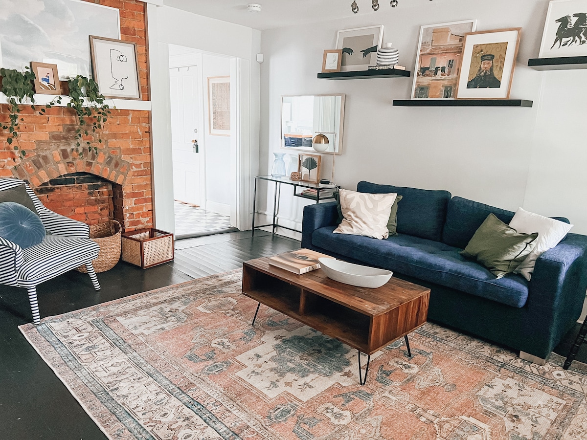 A cozy living area features a blue sofa complemented by decorative pillows. A wooden coffee table sits in front, enhancing the space. An area rug adds warmth, while a stylish chair and a brick fireplace add character. Shelves display curated décor, contributing to the inviting atmosphere.