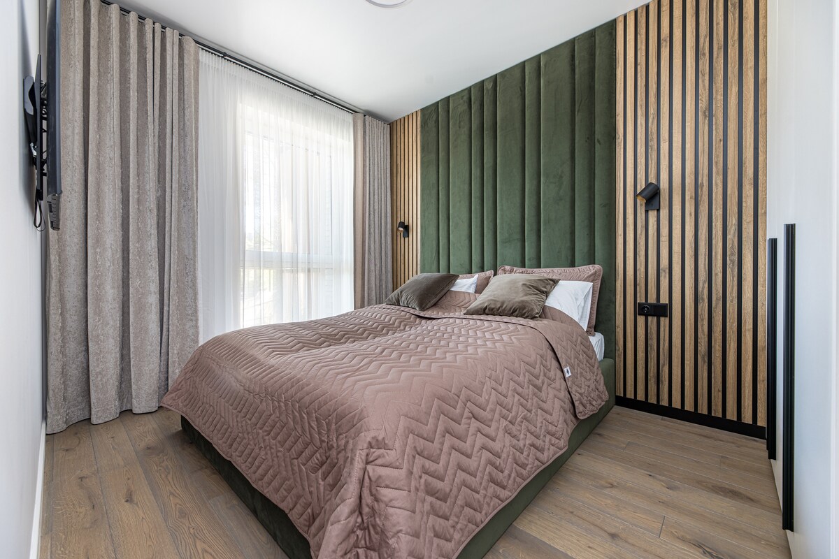 A tranquil bedroom features a spacious bed adorned with a textured, earthy-toned quilt. The walls are accented with wood paneling in varying tones, and light drapes frame the windows, allowing natural light to brighten the room.