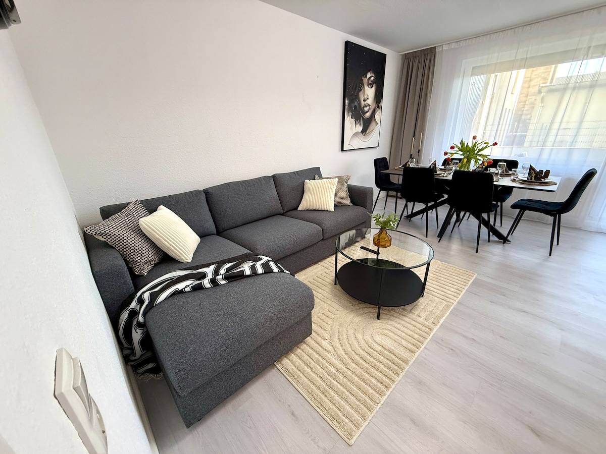 A modern living area features a spacious sectional sofa draped with a patterned throw. A round coffee table sits atop a textured area rug. In the background, a dining set with black chairs is positioned near large windows that allow natural light to fill the space.