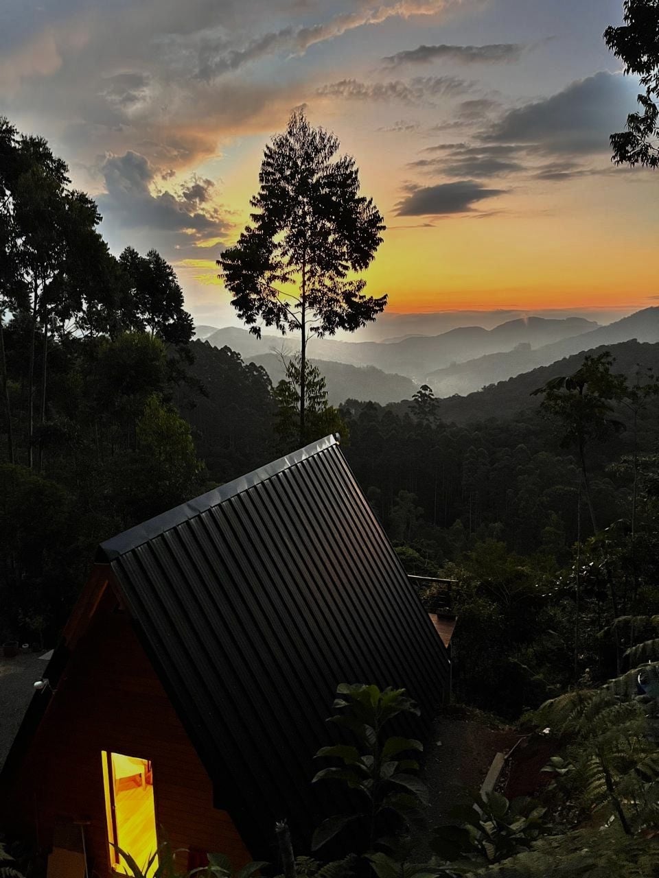 A wooden cabin with a sloped roof is positioned on a hillside, framed by lush greenery. A warm glow emanates from the cabin's window, contrasting with the serene twilight sky. Distant mountains roll gently in the background under an orange and purple sunset.