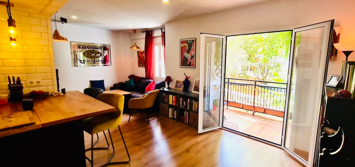 An open living area is visible, featuring a wooden countertop with bar stools, and a bookshelf filled with books and decor. Large glass doors lead to a balcony overlooking greenery, while cozy seating is arranged with colorful cushions. Natural light fills the space.