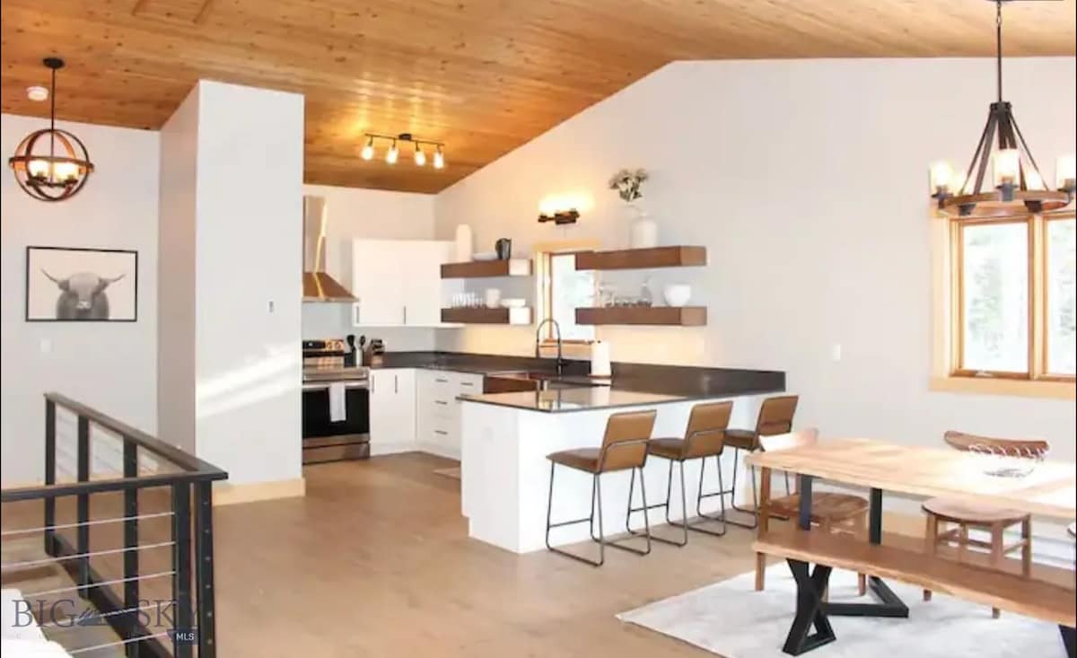 An open-concept kitchen and dining area is showcased, featuring wooden ceilings and light walls. A kitchen island with seating for three is visible alongside a long dining table. Natural light fills the space through large windows, highlighting the modern appliances and decor.