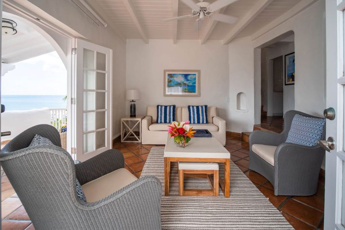 A bright and airy living space features white walls and a ceiling fan, with two cozy armchairs and a light-colored couch arranged around a coffee table. A view of the beach is visible through glass doors, while colorful flowers rest on the table.