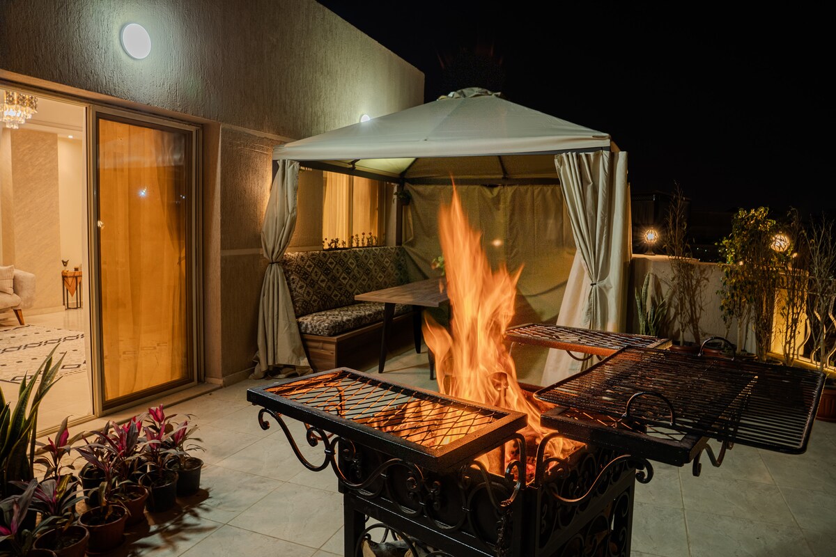 A covered outdoor lounge area is highlighted by a fire pit with flames flickering. Surrounding the seating area, potted plants add greenery, while a spacious table and benches provide ample seating. Soft lighting enhances the ambiance, creating a cozy atmosphere for gatherings.