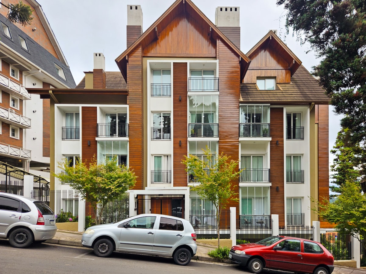 The exterior of a multi-story building is shown, featuring a combination of wooden and light-colored facades. Balconies with glass railings are visible on each level, framed by greenery from nearby trees. Vehicles are parked in front, providing a sense of community and accessibility.