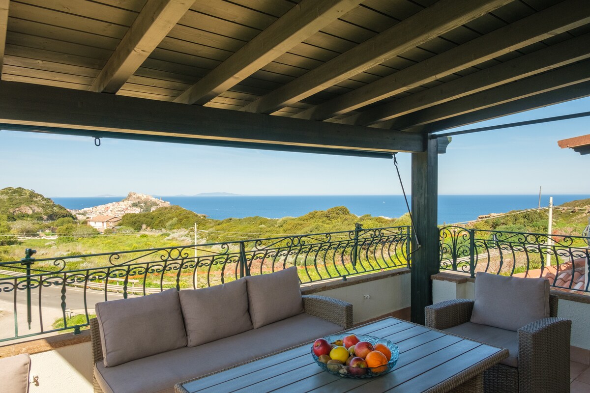 A spacious veranda is furnished with a comfortable seating area comprised of neutral-toned sofas and a wooden table. A colorful bowl of fresh fruits is placed at the center, while a stunning view of the coastline and sea can be seen through the open railing.