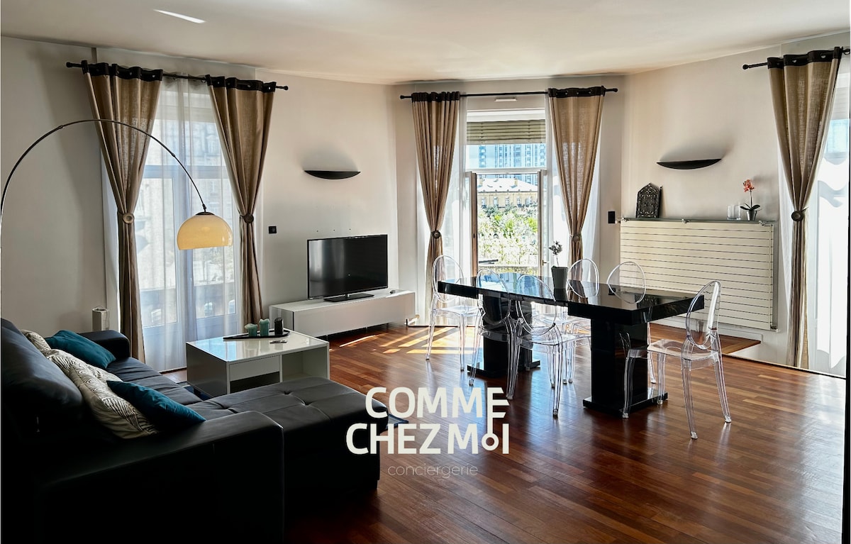A bright living area features a combination of natural light and elegant curtains. A dark sectional sofa is positioned against a wall, complemented by a flat-screen TV and a contemporary dining table with transparent chairs. The balcony doors offer a glimpse of outdoor views.