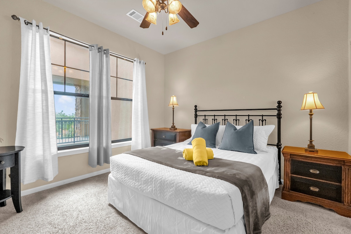 A tranquil bedroom is featured, with a spacious bed adorned with white linens and accent pillows. Two bedside tables with lamps flank the bed, while large windows are framed by light curtains, allowing natural light to fill the room.