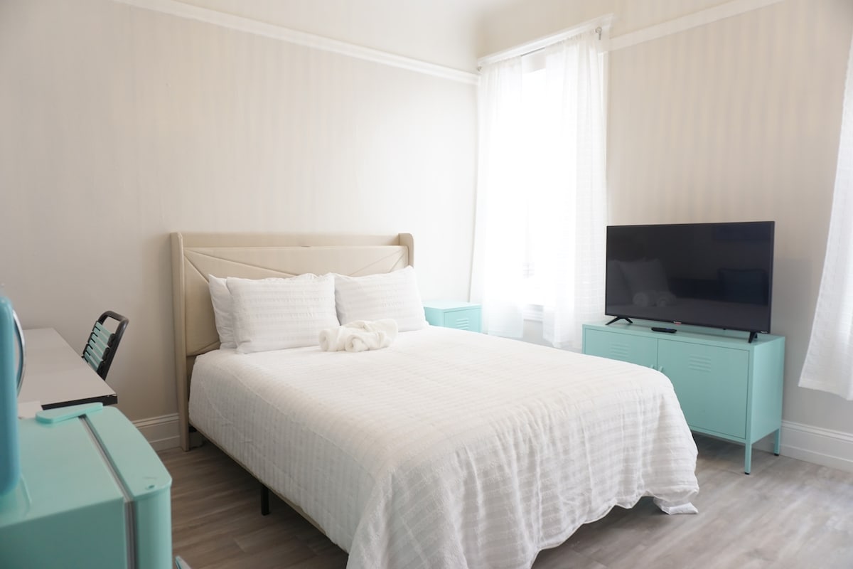 A well-lit bedroom features a spacious bed with white linens, complemented by a cozy towel arranged at the foot. A large television is positioned against a light-colored wall, while teal cabinetry adds a touch of color, creating a serene atmosphere.