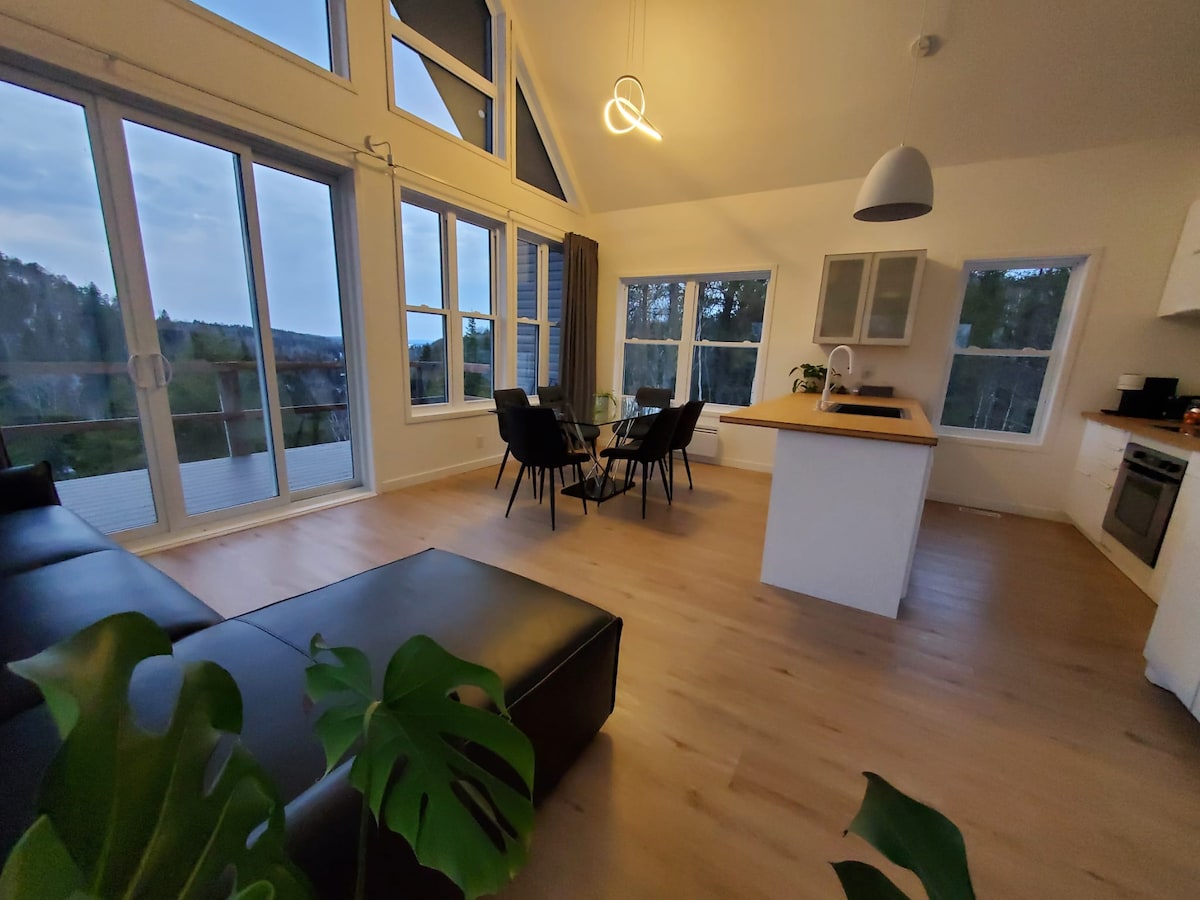 An open-plan living space features a dark sectional sofa and a dining area with four black chairs surrounding a wooden table. Large windows fill the room with natural light, offering views of the surrounding nature. Modern kitchen appliances are visible, enhancing functionality.