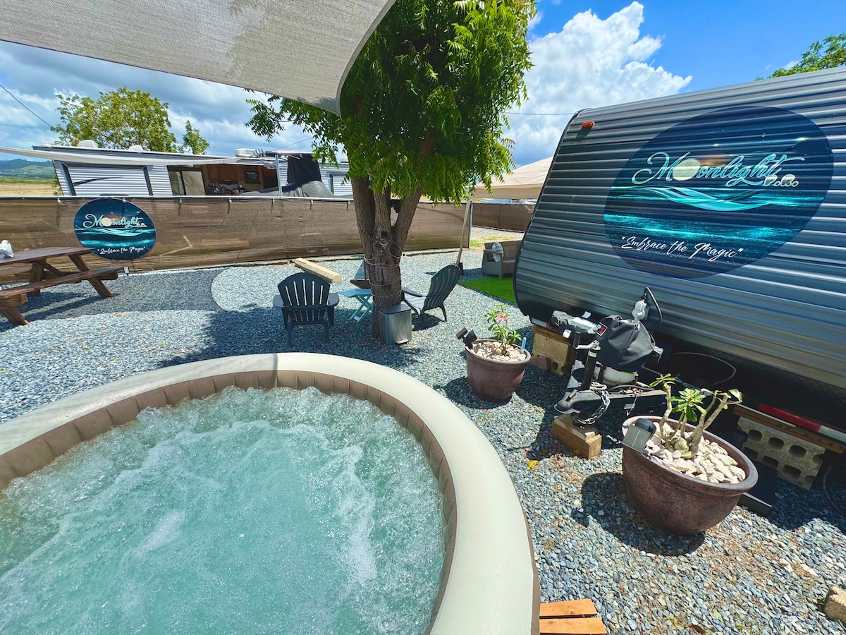 A private jacuzzi is surrounded by gravel and planted pots, creating a relaxing outdoor space. Nearby, two black Adirondack chairs add seating, while the RV looms in the background, partially shaded by a canopy, with a picnic table visible to the side.