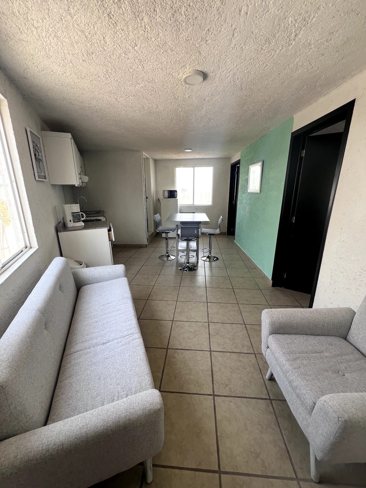 The interior space features a light-colored sofa and a cozy armchair, creating a comfortable seating area. A small dining table with chairs is positioned near a bright window, while functional kitchen elements are visible in the background. Neutral-toned walls and tiled flooring enhance the open layout.