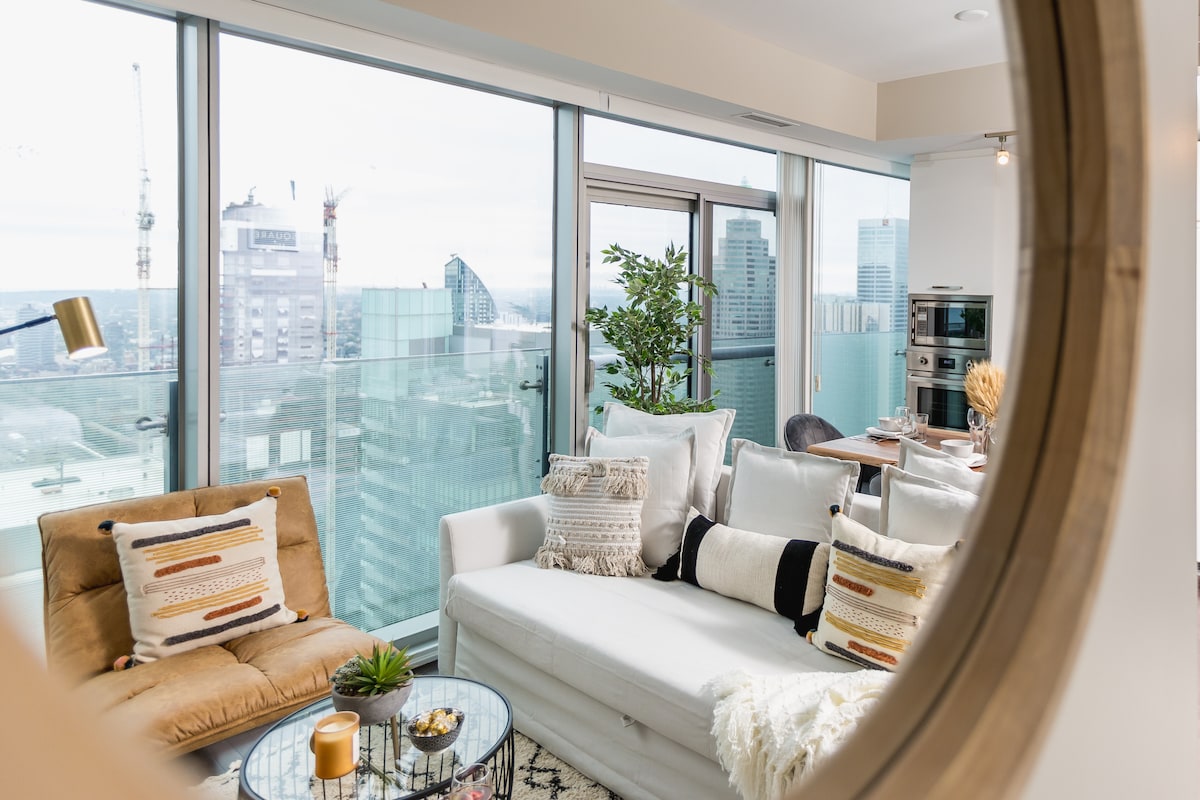 A modern living area features a white sofa and two brown chairs, arranged around a glass coffee table. Large windows offer a view of the city skyline, while a potted plant brings a touch of greenery indoors.