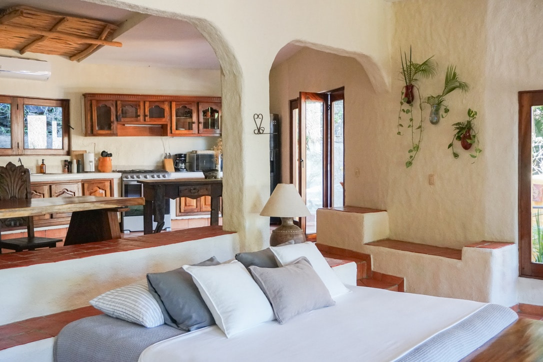A spacious sleeping area features a large bed with multiple pillows arranged neatly. The room includes rustic wooden furniture and an open kitchen area, accented by vibrant plants on the wall. Natural light enters through two adjoining windows, highlighting the warm color palette.
