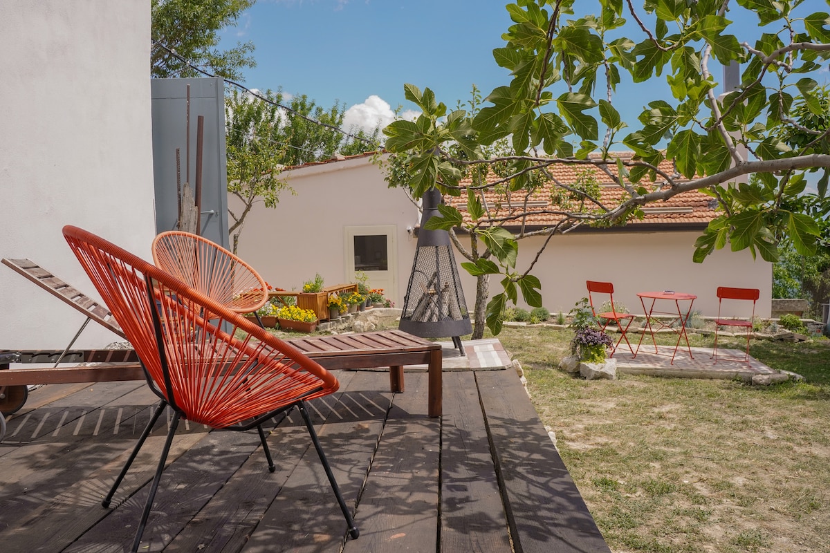 Outdoor seating areas are present, featuring red chairs and a wooden lounger on a deck. The space is shaded by a tree, with a view of the adjoining property. A small dining table with red chairs is visible in the background, surrounded by garden plants.