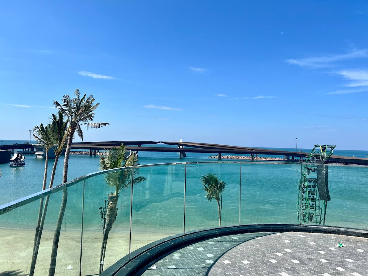 A clear view of the ocean and a bridge is framed by a glass railing. Palm trees are positioned near the water's edge, while a calm sea reflects the blue sky. The image captures the serene atmosphere of beachside living.