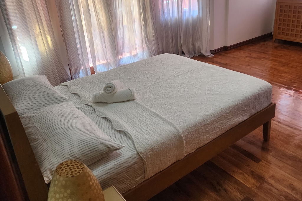 A spacious master bedroom features a king-size bed dressed in soft white linens, accompanied by a rolled towel. Natural light filters through sheer curtains, illuminating the polished wooden floor. The room exudes a tranquil ambiance suitable for relaxation.