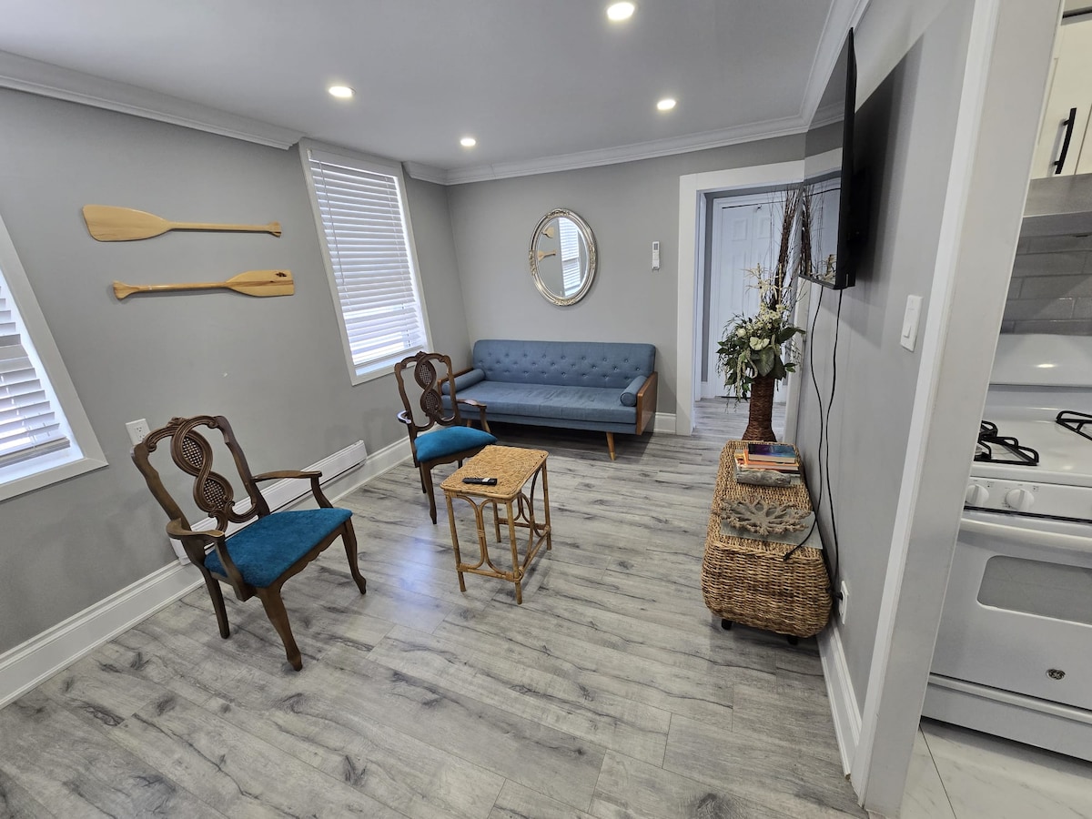 An inviting living area features a blue sofa and two turquoise chairs arranged around a small wooden table. Natural light filters through the windows, highlighting the light wood flooring. A round mirror and decorative elements add to the space's charm.