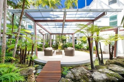 An outdoor seating area is framed by a transparent roof and surrounded by lush greenery. Two comfortable chairs are arranged around a small table on a circular deck, with a wooden walkway leading through thoughtfully landscaped stones and plants.